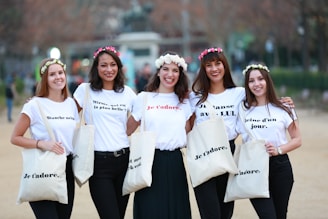 Five people stand together outdoors, each wearing a white shirt with text in French and holding white tote bags printed with 'Je t'adore'. They are adorned with flower crowns and smile at the camera, conveying a sense of friendship and happiness. The background is slightly blurred, suggesting a park or public space.