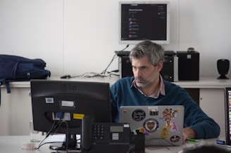 A man is seated at a desk using a laptop adorned with various stickers. He appears focused, likely working on a task. In front of him sits a phone and a computer monitor. The background shows a backpack and some electronic equipment, including a stereo system and a wall-mounted screen displaying an application.