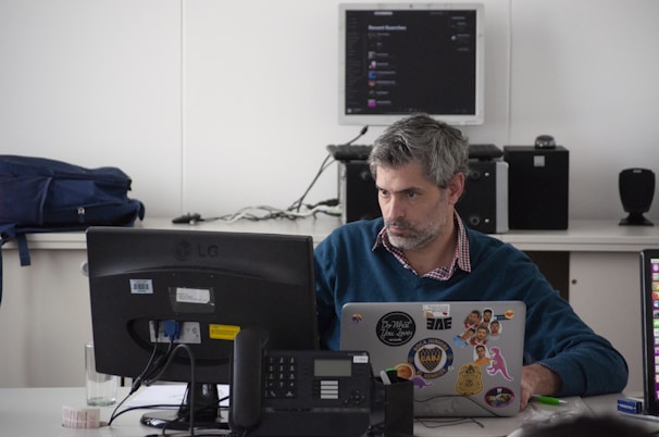 A man is seated at a desk using a laptop adorned with various stickers. He appears focused, likely working on a task. In front of him sits a phone and a computer monitor. The background shows a backpack and some electronic equipment, including a stereo system and a wall-mounted screen displaying an application.