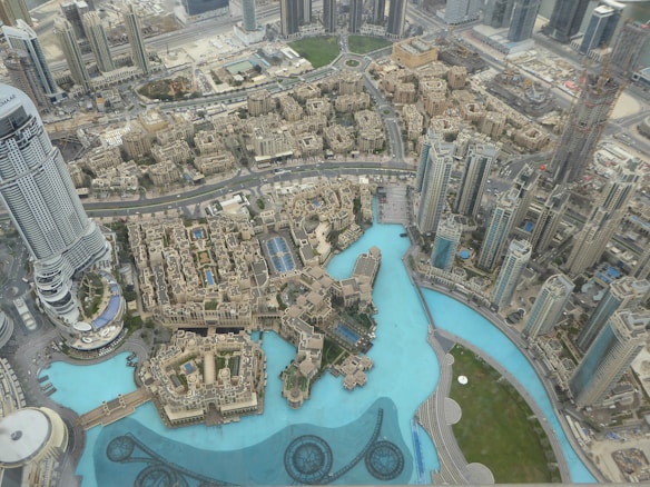 An expansive aerial view of a modern cityscape featuring a mix of skyscrapers and smaller buildings. A large, striking blue pool or artificial lake is surrounded by high-rise architectural structures. Urban density is evident, with a network of roads and greenery visible amongst the buildings.