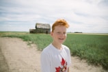 A young person with red hair stands in the foreground of a rural setting, wearing a white shirt with a maple leaf design. Behind them, a dirt path leads to an old, weathered barn surrounded by green fields under a cloudy sky.