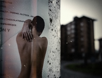 An outdoor scene displaying a poster pasted on a cylindrical surface, featuring a black and white image of a person viewed from the back with their arm reaching over their shoulder. In the background, there is a slightly blurred view of tall apartment buildings against a grey sky, creating an urban atmosphere.