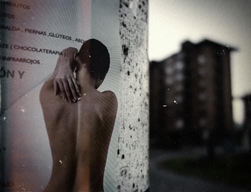 An outdoor scene displaying a poster pasted on a cylindrical surface, featuring a black and white image of a person viewed from the back with their arm reaching over their shoulder. In the background, there is a slightly blurred view of tall apartment buildings against a grey sky, creating an urban atmosphere.
