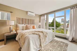A modern, well-lit bedroom featuring a large bed with multiple pillows and a textured throw blanket. The room includes nightstands with lamps on either side of the bed, a stylish headboard with vertical stripes, and a large window offering a view of the city and greenery. The walls are painted in a neutral tone, complementing the soft color palette of the bedding and decor.