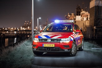 A red emergency response vehicle with blue and white stripes is parked on a paved path at nighttime. The vehicle has a Dutch license plate and 'Rijkswaterstaat' markings. There are headlights on, illuminated streetlights, and blurred industrial structures in the background.