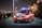 A red emergency response vehicle with blue and white stripes is parked on a paved path at nighttime. The vehicle has a Dutch license plate and 'Rijkswaterstaat' markings. There are headlights on, illuminated streetlights, and blurred industrial structures in the background.