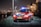 A red emergency response vehicle with blue and white stripes is parked on a paved path at nighttime. The vehicle has a Dutch license plate and 'Rijkswaterstaat' markings. There are headlights on, illuminated streetlights, and blurred industrial structures in the background.