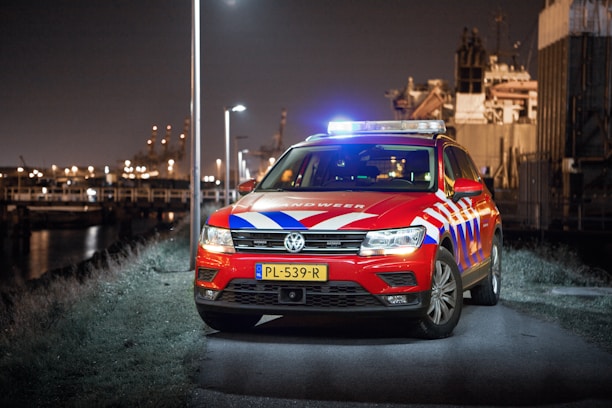 A red emergency response vehicle with blue and white stripes is parked on a paved path at nighttime. The vehicle has a Dutch license plate and 'Rijkswaterstaat' markings. There are headlights on, illuminated streetlights, and blurred industrial structures in the background.