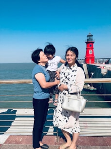 A family scene by a waterfront, featuring two adults and a child. The older adult is holding the child who is leaning towards her, while the younger adult stands beside them holding a white handbag. Behind them, a vivid red lighthouse stands against a clear blue sky.
