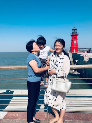 A family scene by a waterfront, featuring two adults and a child. The older adult is holding the child who is leaning towards her, while the younger adult stands beside them holding a white handbag. Behind them, a vivid red lighthouse stands against a clear blue sky.
