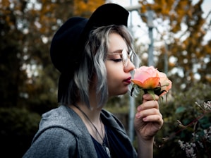 A person with silver hair wearing a black hat and glasses gently smells a peach-colored rose. The background is blurred with greenery and hints of autumn foliage.