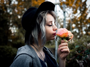 A person with silver hair wearing a black hat and glasses gently smells a peach-colored rose. The background is blurred with greenery and hints of autumn foliage.