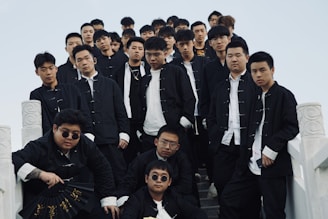 A group of young men wearing matching black jackets and white shirts stand on a set of stone stairs. They appear confident and are looking directly at the camera. One individual at the front holds a black fan with gold inscriptions.