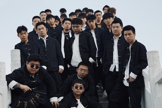 A group of young men wearing matching black jackets and white shirts stand on a set of stone stairs. They appear confident and are looking directly at the camera. One individual at the front holds a black fan with gold inscriptions.