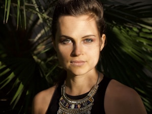 A woman with fair skin and brown hair is looking directly at the viewer. She is wearing a dark top and a decorative necklace with beads and gemstones. The background consists of green palm leaves, casting shadows across her face, giving a natural and serene ambiance.