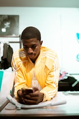 A person is leaning on a table, focusing intently on a smartphone in their hands. They wear a bright yellow track jacket with white stripes and a pendant necklace. Shopping bags and clothing racks are visible in the background, suggesting a retail setting.