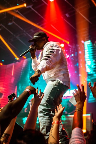 A performer wearing a white jacket and black cap, holding a microphone, is energetically engaging with a lively crowd. The stage is illuminated with vibrant lights, including red, orange, and blue hues, creating a dynamic atmosphere. Audience members are reaching out with their hands, some holding phones to capture the moment.