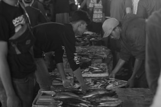 Two men are seen in a market setting, both bent over a table filled with various types of fish. The scene appears busy with other people in the background, indicating an active market environment.