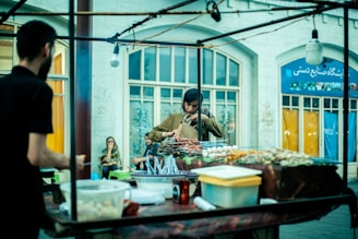 A street vendor stall with a variety of food items on display. A person is standing behind the counter, preparing or arranging food. The background features a building with large windows and people sitting nearby, potentially customers.