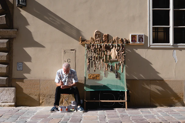 An elderly man is sitting on a small stool in front of a beige wall, carving a piece of wood. He is surrounded by various wooden carvings hanging on a display board, and there are two small religious images beside the display. The setting appears to be a cobblestone street in a sunny, urban environment.