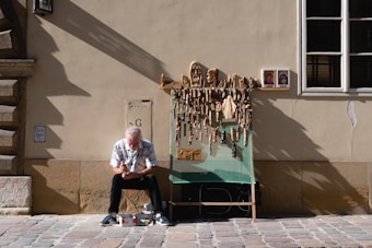 An elderly man is sitting on a small stool in front of a beige wall, carving a piece of wood. He is surrounded by various wooden carvings hanging on a display board, and there are two small religious images beside the display. The setting appears to be a cobblestone street in a sunny, urban environment.