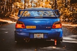 A blue sports car with a large spoiler is parked on an autumn forest road. The car has a 'Student Driver' sticker and a Georgia license plate. The surrounding trees display vibrant orange and yellow fall foliage.