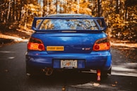 A blue sports car with a large spoiler is parked on an autumn forest road. The car has a 'Student Driver' sticker and a Georgia license plate. The surrounding trees display vibrant orange and yellow fall foliage.