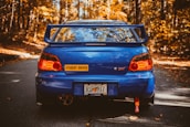 A blue sports car with a large spoiler is parked on an autumn forest road. The car has a 'Student Driver' sticker and a Georgia license plate. The surrounding trees display vibrant orange and yellow fall foliage.