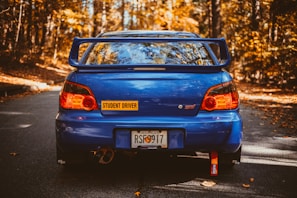 A blue sports car with a large spoiler is parked on an autumn forest road. The car has a 'Student Driver' sticker and a Georgia license plate. The surrounding trees display vibrant orange and yellow fall foliage.