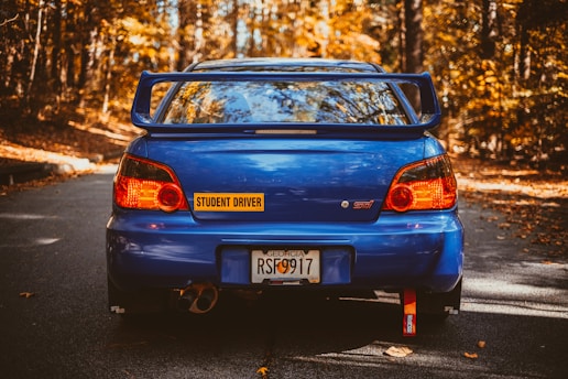 A blue sports car with a large spoiler is parked on an autumn forest road. The car has a 'Student Driver' sticker and a Georgia license plate. The surrounding trees display vibrant orange and yellow fall foliage.