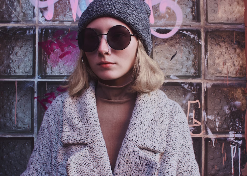 woman wearing white and gray coat and sunglasses leaning against the wall