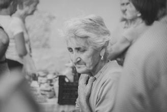 An elderly woman with short gray hair stands amidst a group of people. Her expression is thoughtful and slightly somber. The setting appears to be casual and social, with people in the background engaged in conversation. The image is in black and white, adding a nostalgic feel.