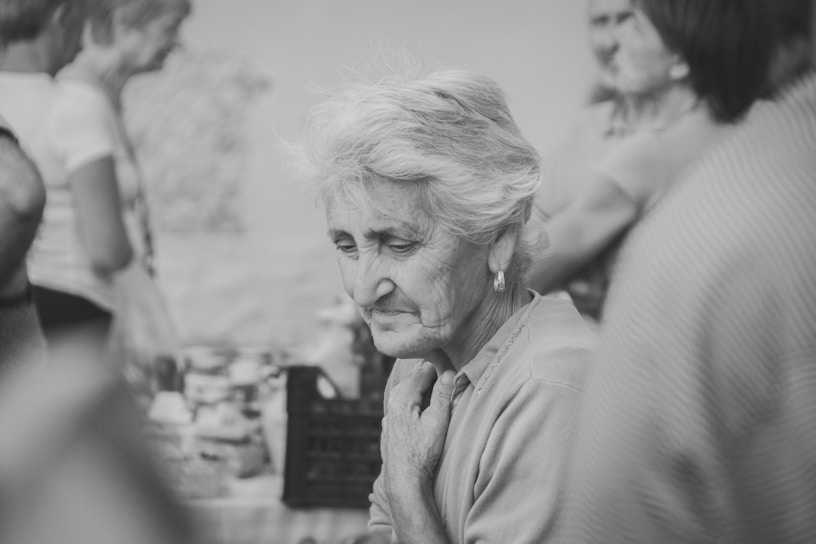 An elderly woman with short gray hair stands amidst a group of people. Her expression is thoughtful and slightly somber. The setting appears to be casual and social, with people in the background engaged in conversation. The image is in black and white, adding a nostalgic feel.