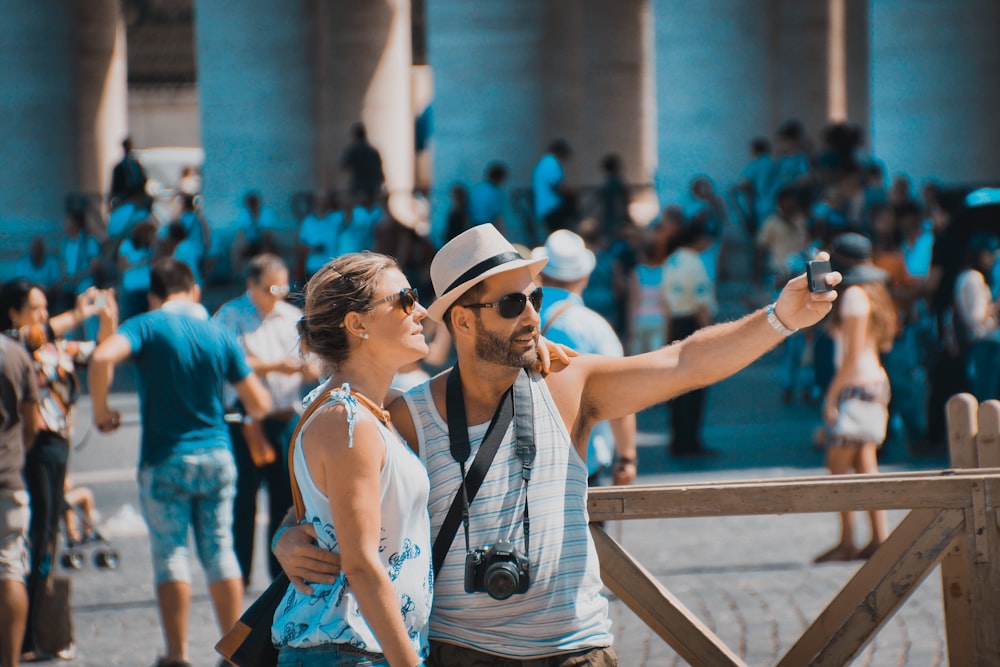 Casal de turistas, um homem e uma mulher, tirando fotos em frente a um ponto turístico. O homem tem uma câmera pendurada no pescoço, óculos escuro, chapéu e tira a foto com o celular, enquanto a mulher utiliza um óculos escura e se abraça a ele.
