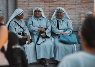 Three nuns dressed in blue and white habits are sitting together on stone steps, engaging in conversation. They are wearing glasses and hold bags and rosaries. The background consists of a brick wall, and other people can be seen blurred in the foreground and background.