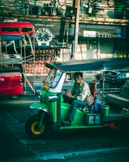 A green tuk-tuk with a driver sitting inside, parked on a street. Behind it, a busy city scene with colorful shops and signs, and a red vehicle partially visible. Overhead wires crisscross above the scene.