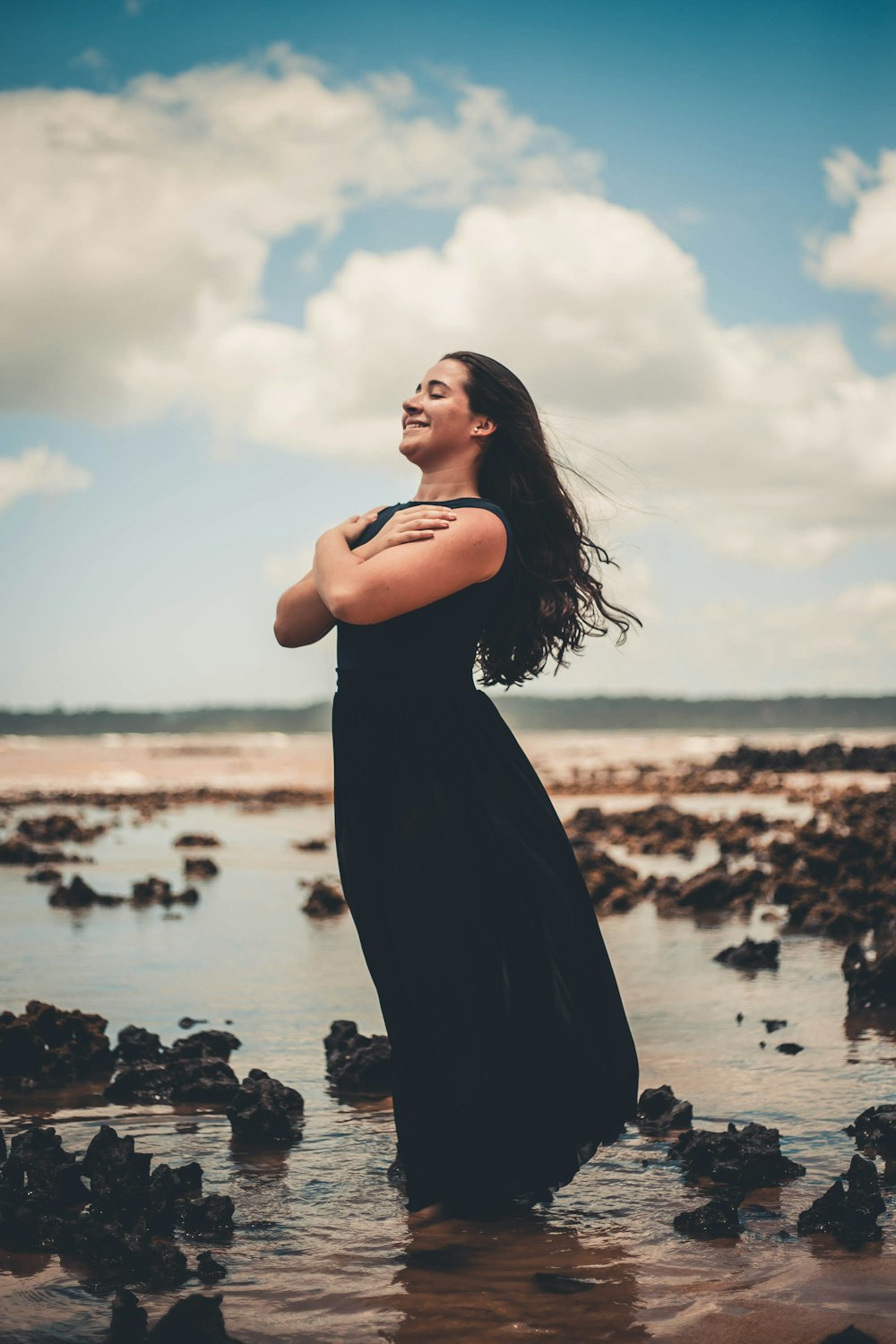 smiling woman wearing black sleeveless dress standing on body of water under white and blue sky