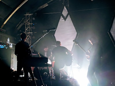 A band is performing on stage with instruments including a guitar, keyboard, and drums. The lighting is dim, creating a dramatic silhouette effect on the musicians. The background features geometric shapes and spotlights, with a haze from stage lighting.