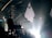 A band is performing on stage with instruments including a guitar, keyboard, and drums. The lighting is dim, creating a dramatic silhouette effect on the musicians. The background features geometric shapes and spotlights, with a haze from stage lighting.