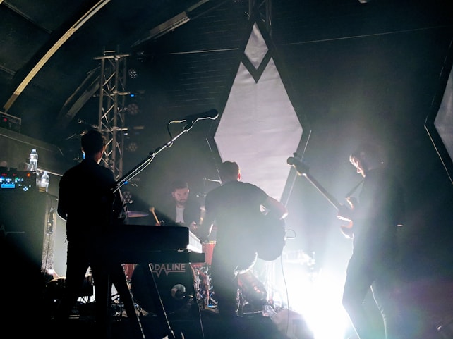 A band is performing on stage with instruments including a guitar, keyboard, and drums. The lighting is dim, creating a dramatic silhouette effect on the musicians. The background features geometric shapes and spotlights, with a haze from stage lighting.