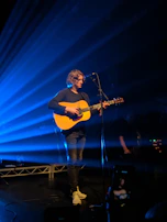 A musician with long hair is performing on stage, playing an acoustic guitar and standing in front of a microphone. Beams of blue light radiate from the background, creating a dynamic and moody atmosphere. The musician is wearing casual clothes, including a dark top and light sneakers.