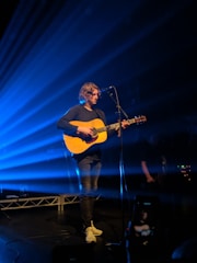 A musician with long hair is performing on stage, playing an acoustic guitar and standing in front of a microphone. Beams of blue light radiate from the background, creating a dynamic and moody atmosphere. The musician is wearing casual clothes, including a dark top and light sneakers.