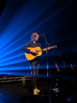 A musician with long hair is performing on stage, playing an acoustic guitar and standing in front of a microphone. Beams of blue light radiate from the background, creating a dynamic and moody atmosphere. The musician is wearing casual clothes, including a dark top and light sneakers.