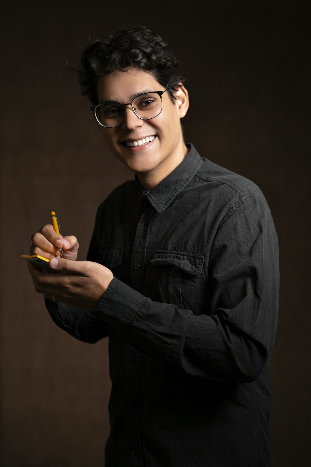 Hombre sonriente con chaqueta de mezclilla negra sosteniendo lápiz y nota adhesiva mientras está de pie