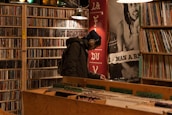 A person wearing headphones and a beanie is browsing records in a dimly lit music store. The store has shelves filled with CDs and vinyl records. There is a large poster in the background with a musician's image. The person is bent over, closely inspecting the records.