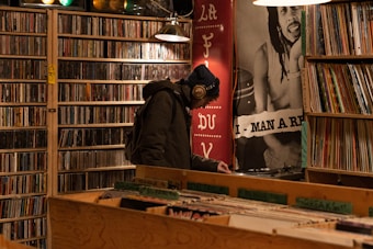 A person wearing headphones and a beanie is browsing records in a dimly lit music store. The store has shelves filled with CDs and vinyl records. There is a large poster in the background with a musician's image. The person is bent over, closely inspecting the records.