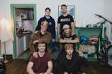 A group of six young individuals is gathered in a cozy room. The room features a mix of furniture and personal items, including a bicycle, shelves with assorted items, and a corner with a kitchen visible. Some wear casual attire with t-shirts, hoodies, and denim. One person is seated on a stool, while others pose at different heights.
