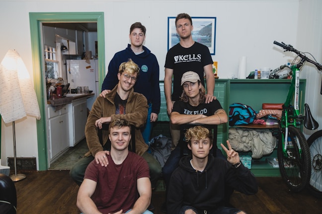 A group of six young individuals is gathered in a cozy room. The room features a mix of furniture and personal items, including a bicycle, shelves with assorted items, and a corner with a kitchen visible. Some wear casual attire with t-shirts, hoodies, and denim. One person is seated on a stool, while others pose at different heights.