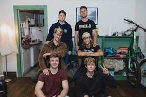 A group of six young individuals is gathered in a cozy room. The room features a mix of furniture and personal items, including a bicycle, shelves with assorted items, and a corner with a kitchen visible. Some wear casual attire with t-shirts, hoodies, and denim. One person is seated on a stool, while others pose at different heights.