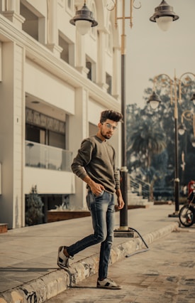 A fashionable young man with styled hair and sunglasses stands casually on a sidewalk next to a modern building. He is wearing a green sweater, jeans, and sneakers. The scene includes street lamps and a motorcycle parked in the background.